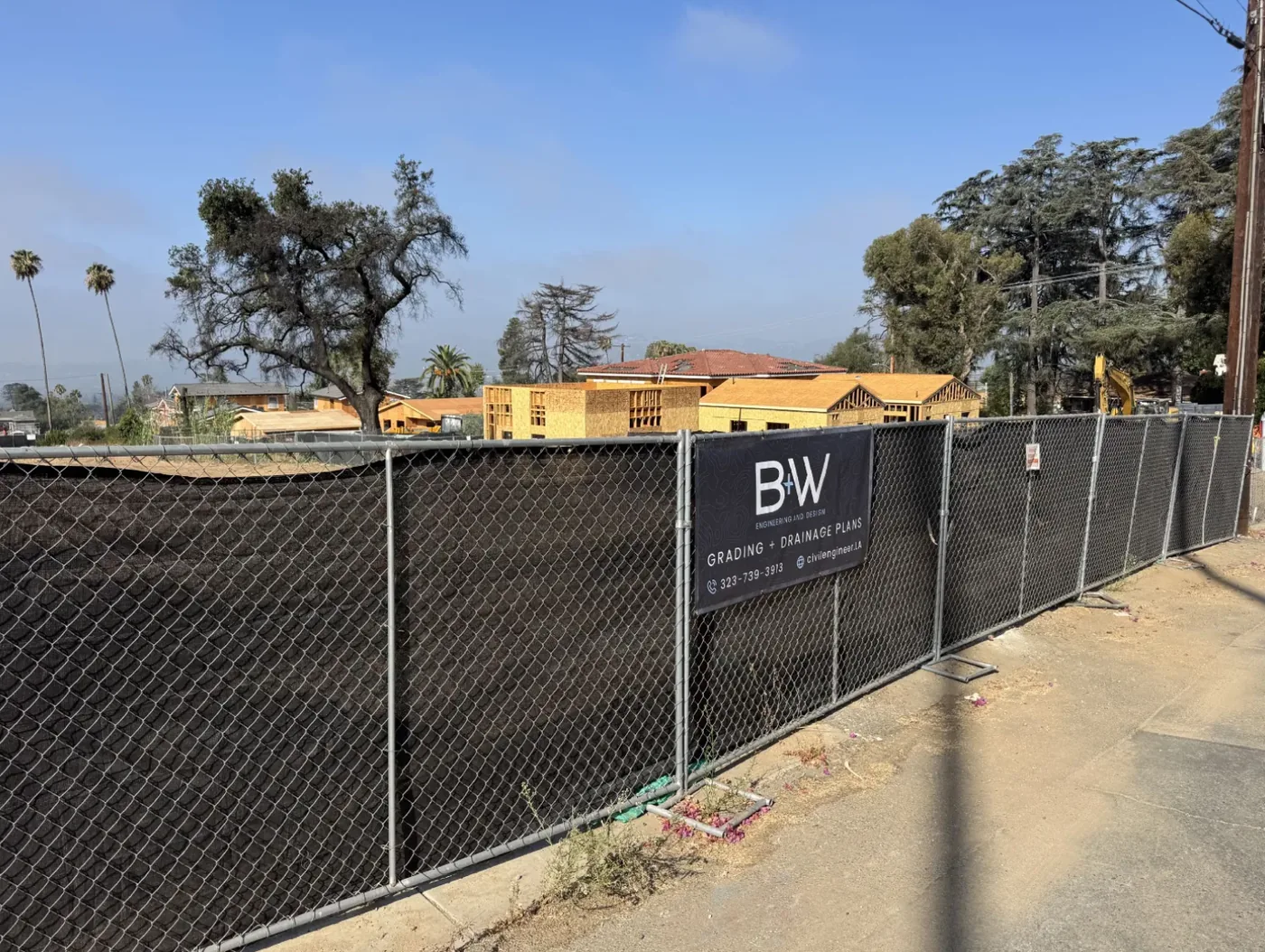 Long run of temporary chain link fencing along an Altadena sidewalk with the B+W banner mounted mid-run, framed homes under construction beyond it